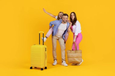 Sarı bavullu oyuncu aile sarı arka planda stüdyoda poz veriyor. Babası geri dönen bir kız, elinde çantasıyla gülümseyen bir anne, eğlenceli bir tatil anlayışı.
