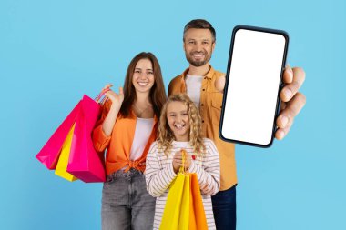 Mutlu aile alışverişleri stüdyoda. Ebeveynler ve kızları renkli çantalar taşıyorlar ve reklam için boş ekranlı akıllı telefonları veya online alışveriş konseptini gösteriyorlar.