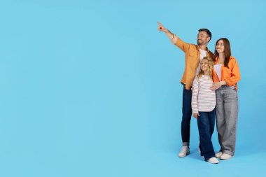 Gülümseyen aile mavi arka planda stüdyoda yan yana dururken fotokopi alanını işaret ediyor. Baba ileri görüşlü, vizyonu sembolize eden takım çalışması ve pozitif aile geleceğini işaret ediyor.