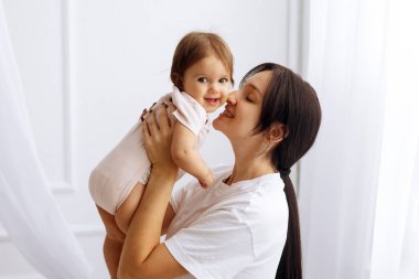 Mutlu anne evde gülümserken bebeğini kucağına alıp öpüyor. Parlak doğal ışık, sıcaklığı ve sevgi dolu aile bağını arttırır.