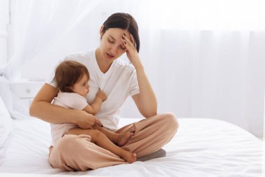 Yorgun anne kucağında bebekle yatakta oturuyor, bitkin görünüyor. Ebeveynlik ve duygusal yorgunluğun günlük zorluklarını gösteren gerçekçi aile sahnesi, kopyalama alanı