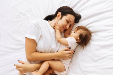 Anne beyaz yatakta uzanıyor ve bebeğini sıkıca kucaklıyor. Ebeveyn ve çocuk arasındaki samimiyet, koruma, sevgi ve barışçıl bağı gösteren güzel bir aile sahnesi.