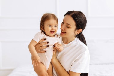 Gülümseyen anne bebeğini kucağında tutuyor ve sevgiyle bakıyor. Aile hayatında şefkat, neşe ve duygusal bağlantıyı ifade eden parlak kapalı mekan sahnesi