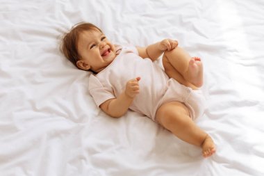 Gülen bebek beyaz yatakta uzanıyor, neşeyle ayaklarıyla oynuyor. Mutlu çocukluk konsepti evde masumiyet, özgürlük ve şen şakrak ruhu gösteriyor