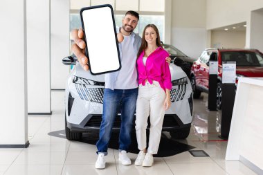 Gülümseyen genç çift arabanın yanında duruyor ve ellerinde boş ekranlı akıllı telefon, yaşam tarzı konsepti teknoloji, ulaşım, yeni araba sahibi ve modern iş