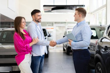 Gülümseyen araba satıcısı bayilik merkezinde müşterilerle tokalaşıyor. Modern otomobil galerisinde başarılı bir araba anlaşması ve olumlu bir anlaşma anı