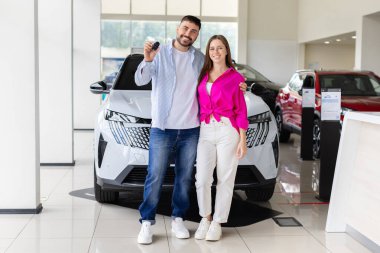 Yeni arabanın önünde elinde anahtarlar olan gülümseyen bir çift. Parlak galeride satın alınmış araçlarıyla poz veren gururlu genç alıcılar.