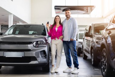 Yeni arabanın yanında duran gülümseyen bir çift. Parlak modern sergi salonunda satın alınmış araçlarla gururla poz veren genç alıcılar.
