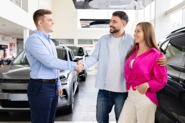 Başarılı bir satın almadan sonra gülümseyen çift araba satıcısıyla tokalaşıyor. Mutlu müşteriler anlaşmayı kapatıyorlar ve galeriye olan güveni onaylıyorlar.