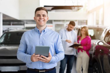 Satış elemanı elinde tabletle, arka planda evrakları kontrol eden mutlu bir araba satıcısı. Otomobil galerisinde kendinden emin ve başarılı bir satış temsilcisi
