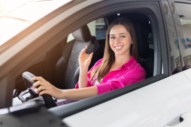 Şoför koltuğunda oturan gülümseyen genç bir kadın. Parlak galeride satın aldıktan sonra yeni bir araçla poz veren mutlu bayan müşteri.