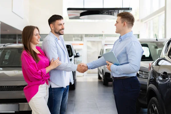 Gülümseyen araba satıcısı bayilik merkezinde müşterilerle tokalaşıyor. Modern otomobil galerisinde başarılı bir araba anlaşması ve olumlu bir anlaşma anı