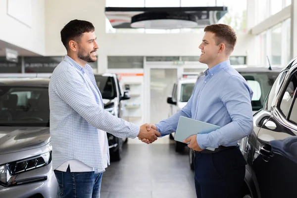 Tebessüm eden müşteri galeride anlaşmadan sonra araba satıcısıyla tokalaşıyor. Modern otomotiv perakendecisinde güven ve başarıyı sembolize eden olumlu iş etkileşimi