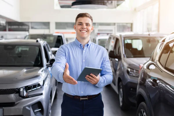 Modern araba galerisinde el sıkışmak için elini uzatan gülümseyen bir satıcı. Güvenilir genç adam müşteriyi selamlıyor, profesyonel araç sunumuna başlamaya hazır.