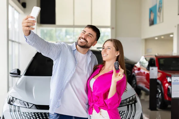 Neşeli çift yeni araba anahtarıyla selfie çekiyor. Gülümseyen adam ve kadın arabalarını galeriden aldıktan sonra mutlu anları yakalıyorlar.