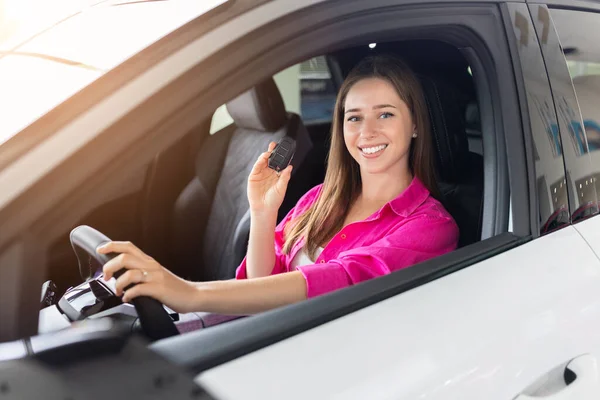 Şoför koltuğunda oturan gülümseyen genç bir kadın. Parlak galeride satın aldıktan sonra yeni bir araçla poz veren mutlu bayan müşteri.