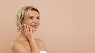 Havluya sarılmış, yanağına dokunan, gülümseyen, cilt bakımını seven, bej renkli stüdyo arka planında duran, gülümseyen olgun bir kadın. Yüksek kalite fotoğraf