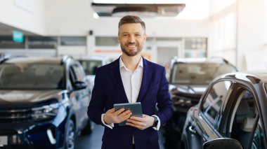 Siyah takım elbiseli ve beyaz gömlekli profesyonel bir araba satıcısı lüks araçlarla dolu modern araba galerisinde duruyor. Elinde dijital tablet tutarken gülümseyen bir adam kameraya bakıyor.
