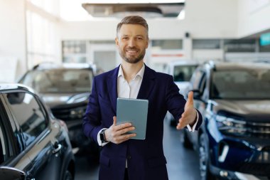 Araba satıcısı dijital tablet tutuyor, el sıkışmak için selamlıyor ve uzatıyor, arka planda modern araçlarla bayilik merkezinde çalışırken içten bir gülümsemeyle gülümsüyor, boşluk kopyalıyor.