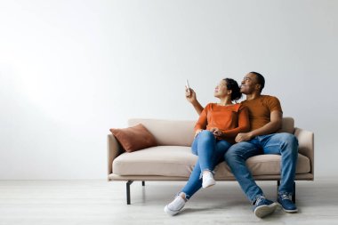 Sistemi ayır. Neşeli genç siyah çift klimayı uzaktan kumandayla açıp beyaz stüdyo arka planında koltukta otururken mutlu Afrikalı Amerikalı erkek ve kadın klima kullanıyor.