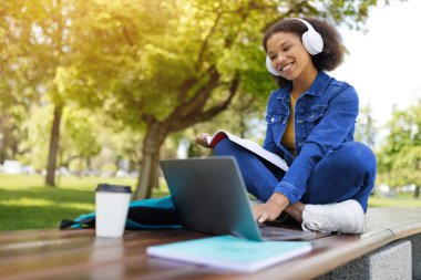 Mutlu siyahi kız öğrenci dışarıda dizüstü bilgisayar ve çalışma kitaplarıyla çalışıyor, Afrikalı Amerikalı kadın kampüste bankta otururken bilgisayar kullanıyor, sınavlara hazırlanıyor, boş yer var.