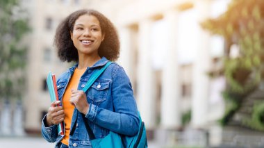 Dışarıda dikilen mutlu, genç siyahi bir kadının portresi çalışma kitapları ve sırt çantasıyla gezen, yan bakan ve gülümseyen, uzay kopyası