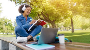 Mesafe öğrenme. Genç siyah kız öğrenci dışarıda dizüstü bilgisayarla çalışıyor bilgisayarını kullanan mutlu Afrikalı Amerikalı kadın ve parkta bankta otururken not defteri tutuyor.