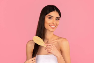 Uzun düz saçlı, gülümseyen ve pembe arka planda nazikçe fırçalayan genç bir kadın. Stüdyo çekimi güzellik rutinini, sağlıklı saç ve kişisel bakım kavramını temsil eder.