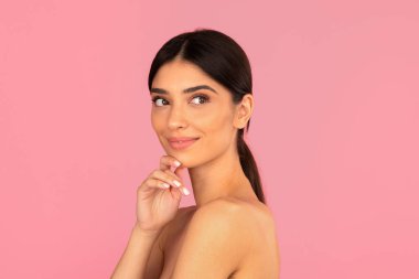 Yumuşak tenli gülümseyen kadın yan bakarken nazikçe çenesine dokunuyor. Özgüveni, güzelliği ve doğal kadınlığı simgeleyen stüdyo portresi.