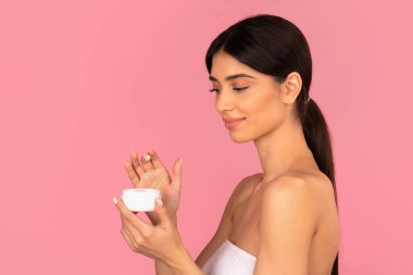 Elinde krema kavanozu olan gülümseyen genç bir kadın ona neşeyle bakıyor, yeni bir ürün yolluyor. Pembe arka plandaki stüdyo portresi günlük cilt bakımını ve kişisel bakım rutinini temsil ediyor.