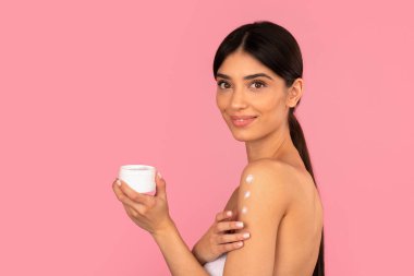 Gülümseyen kadın omzuna yumuşacık bir hareketle nemlendirici krem sürüyor ve kolunda üç nokta oluşturuyor. Vücut bakımını, güzelliğini ve cilt hidrasyonunu gösteren stüdyo portresi