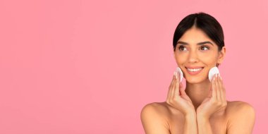 Gülümseyen kadın pembe arka planda pamuk pedlerle yüzünü temizliyor. Günlük cilt bakımını, yüz temizliğini ve tazeliği sembolize eden stüdyo portresi