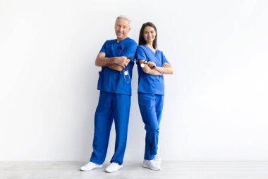 Kıdemli erkek doktor ve mavi önlüklü genç bayan doktor, beyaz arka plan, profesyonel sağlık ekibi konsepti üzerinde güvenle poz veriyorlar.