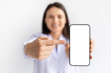 Beyaz önlüklü, gülümseyen ve akıllı telefona işaret eden beyaz arka plan, sağlık teknolojisi ve dijital hizmetler kavramı boş ekranlı kadın doktor.