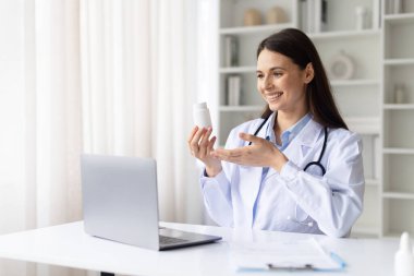 Beyaz önlüklü, elinde ilaç şişesi olan gülümseyen kadın doktor. Parlak klinik ofisinde online danışmanlık sırasında dizüstü bilgisayarla ilaçları açıklıyor.
