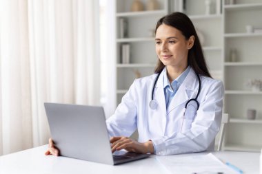 Beyaz önlüklü gülümseyen kadın doktor, parlak klinik ofisinde dizüstü bilgisayarda çalışıyor, profesyonel sağlık uzmanı tıbbi danışmanlık için teknolojiyi kullanıyor.