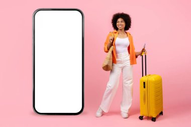 Afro-Amerikalı kadın gezgin. Akıllı telefon modelinin yanında duruyor. Pembe arka planda bavuluyla. Seyahat ve dijital uygulama tanıtım kavramı