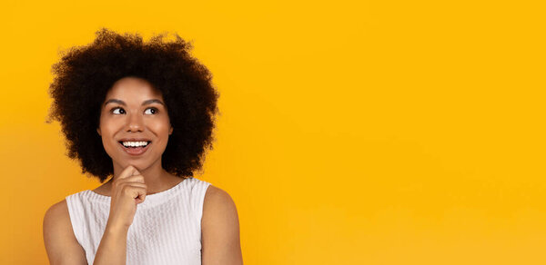 Smiling young African American woman thinking and looking away with hand on chin, isolated on yellow background. Concept of ideas, inspiration, and imagination