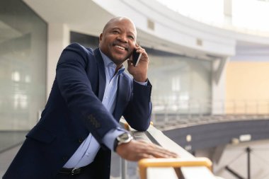 Düşünceli, olgun, Afro-Amerikan iş adamı parmaklıklara yaslanmış, kurnaz bir gülümsemeyle geleceğe bakıyor. Modern bir binadaki hedefleri ve fırsatları yansıtan yönetici