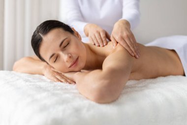 Genç bayan masaj yaptırırken hafifçe gülümsüyor. Sıcak ışık ve yumuşak atmosfer rahatlığı, güzellik bakımını, cilt bakımını ve neşeli bir yaşam tarzını vurgular.