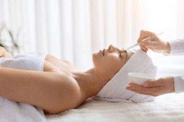 Genç bir kadın modern spa merkezinde yüz maskesi uygulaması alıyor. Terapist ürün uygulamak için fırça kullanır, cilt bakımı ve güzellik bakımını vurgular
