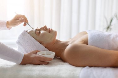 Güzellik uzmanı kaplıcadaki güzel bayana yüz kilinden maske takıyor. Kadın müşteri güzellik hizmetlerinden, cilt bakımından ve huzur dolu yaşam tarzından zevk alıyor.