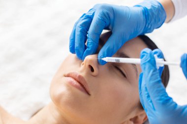 Kozmetik enjeksiyon, kaplıcadaki genç bir kadının burun bölgesine uygulanır. Profesyonel eldivenler, dikkatli baskı ve sakin ifade, temiz kaplıca tedavisi ortamında tipik tıbbi estetiği resmeder.