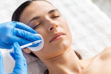 Genç bir kadın pamuk desteğiyle yanak bölgesine estetik enjeksiyon yaptırıyor. Temiz spa seti ve kusursuz eldiven tekniği kozmetik bakım, güzellik geliştirme ve dermatoloji hassasiyetini vurguluyor.