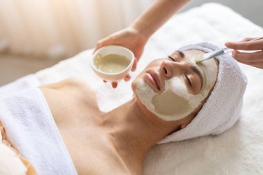 Güzellik uzmanı, kaplıcada genç ve güzel bir bayana kilden maske takıyor. Güzellik terapisti fırçayla cilt bakımı uyguluyor, sağlık ve rahatlama sağlıyor.