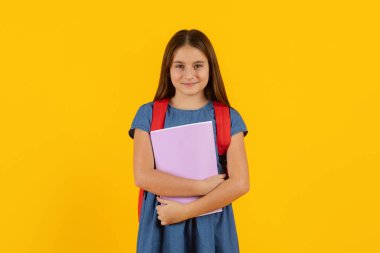 Sırt çantası ve parlak sarı stüdyo arka planında not defteri olan genç bir kız. Öğrenimi, eğitimi ve temiz bir modern okul yaşam tarzını temsil eden kendinden emin bir öğrenci.