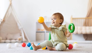 Gülen bebek elinde renkli top ve yüzük oyuncağıyla yerde oturuyor. Parlak ev içi ve dağınık oyuncaklar eğlenceli keşifler ve erken motor beceri geliştirmenin altını çiziyor