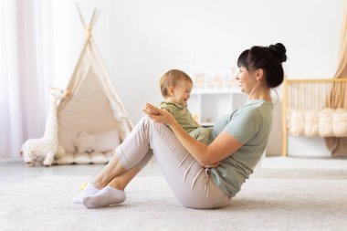 Annem kucağında gülen bebeğiyle yerde oturuyor. Neşeli etkileşim ve yumuşak ev geçmişi aile hayatının sıcaklığını, neşesini ve doğal yakınlığını yansıtıyor.