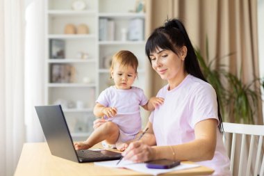 Annesi bebeği masanın yanında otururken notlar yazıyor. Sahne, uzaktan çalışma, planlama, çocuk bakımı ve günlük ev yaşamındaki verimlilik ve aile etkileşimi dengesini yansıtıyor.
