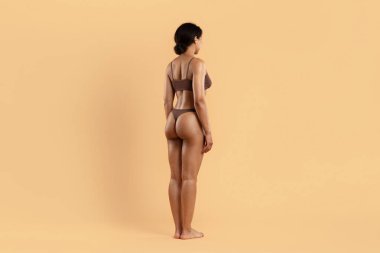 Kahverengi iç çamaşırlı, bej arka planda genç siyahi bir kadın. Renklendirilmiş arka, yontulmuş kalça ve pürüzsüz hatlar güzellik, kilo kaybı ve vücut tonu görselleri için uygun